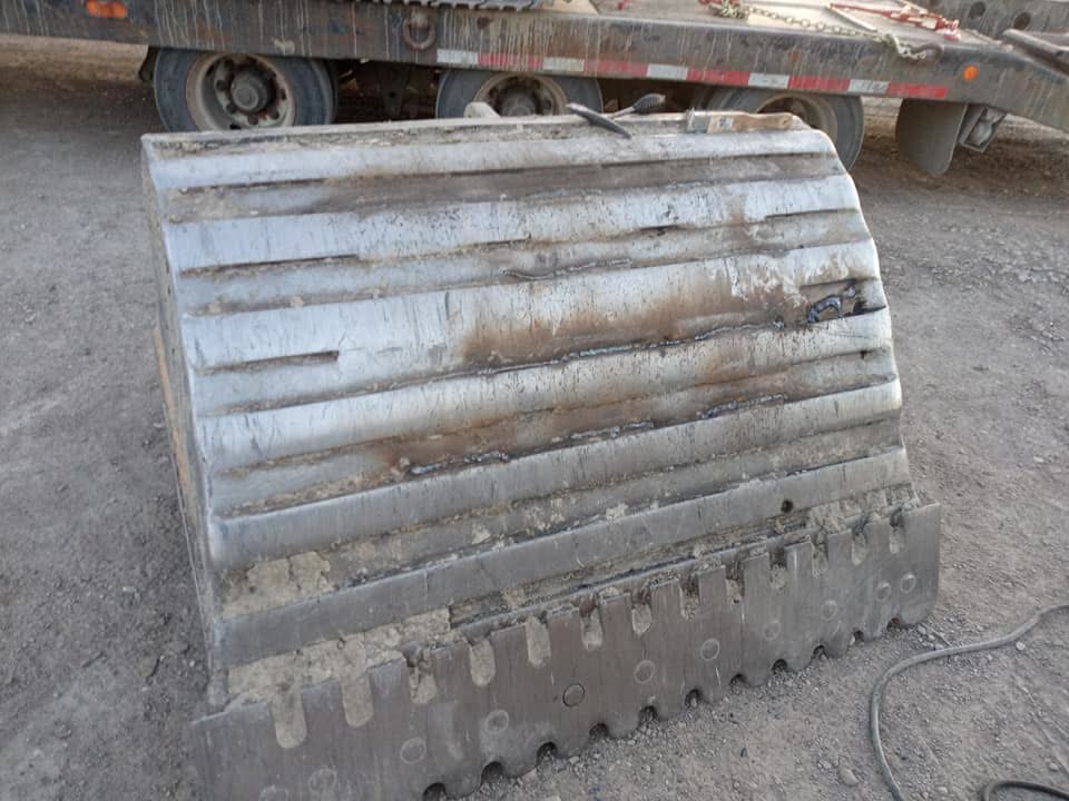 Excavator bucket with weld-repaired ribs and cutting edge sitting in a gravel yard.